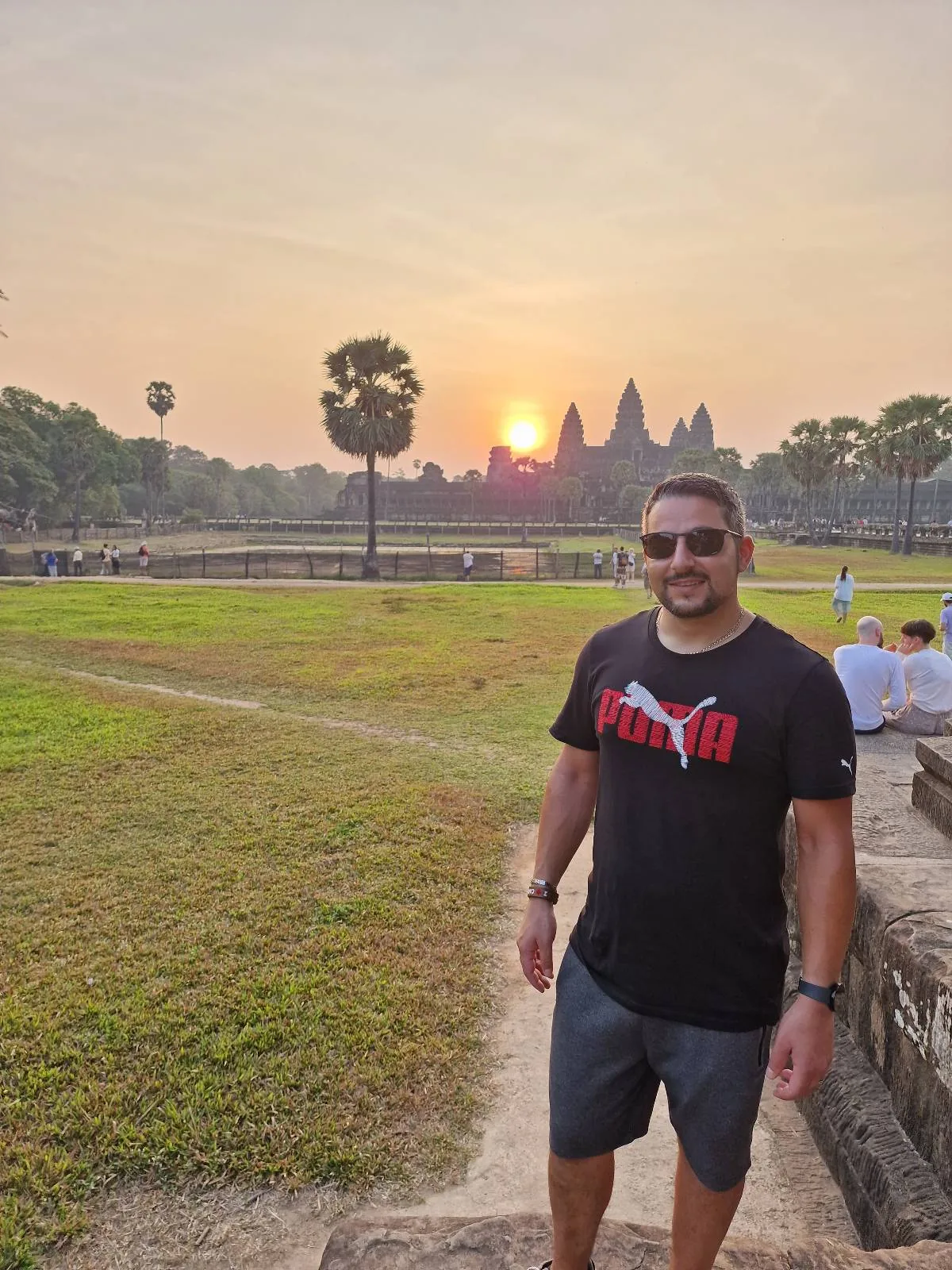 A man wearing sunglasses, a black Puma t-shirt, and shorts stands outdoors in front of Angkor Wat at sunrise, with green grass and trees around and the sun rising behind the temple.