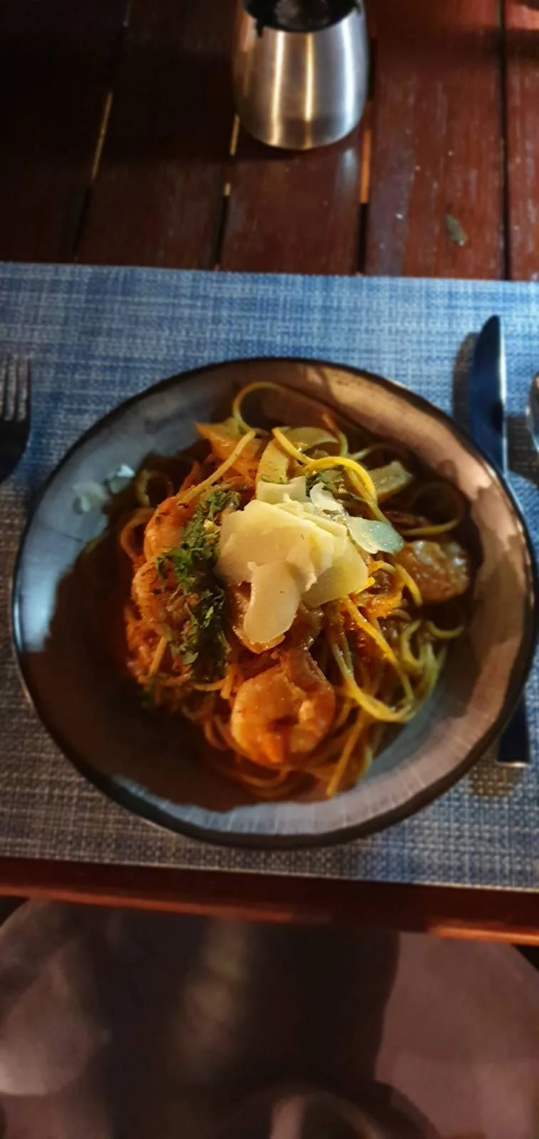 A bowl of pasta garnished with shrimp, sliced vegetables, and herbs, served on a blue placemat with a fork and knife on either side. The dish is topped with shavings, possibly cheese, and is on a wooden table.