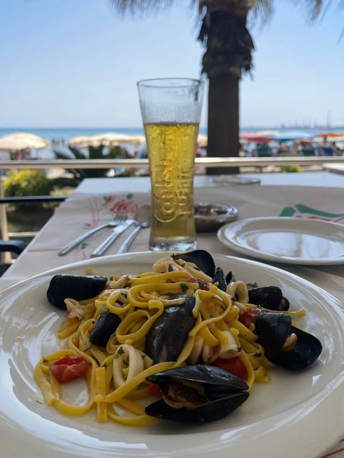 A plate of seafood pasta with mussels sits on a table overlooking a beach with umbrellas and palm trees. A tall glass of beer is next to the plate, with the sea visible in the background.