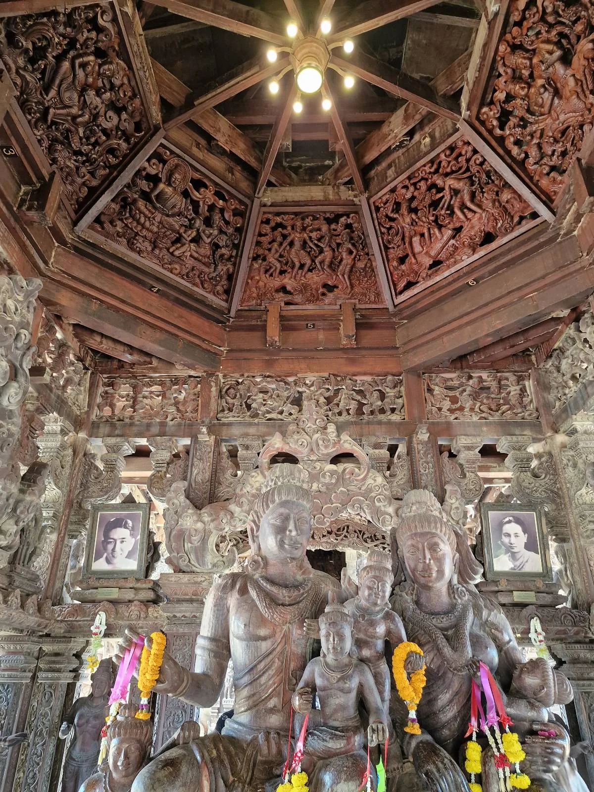A detailed wooden sculpture featuring three figures is set inside an intricately carved wooden temple. The ceiling is adorned with ornate carvings, and framed portraits are visible in the background. Vibrant floral garlands embellish the statues.