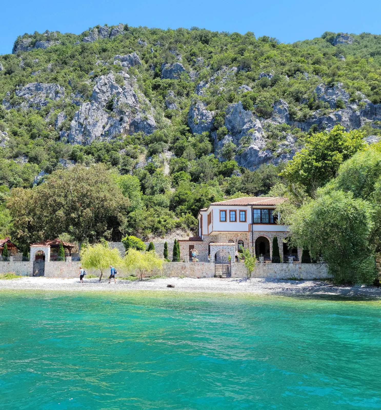 A scenic view of a coastal villa surrounded by lush greenery and rocky hills. The villa has a terracotta roof and large windows. The turquoise sea occupies the foreground, while a small sandy beach lies between the water and the villa.
