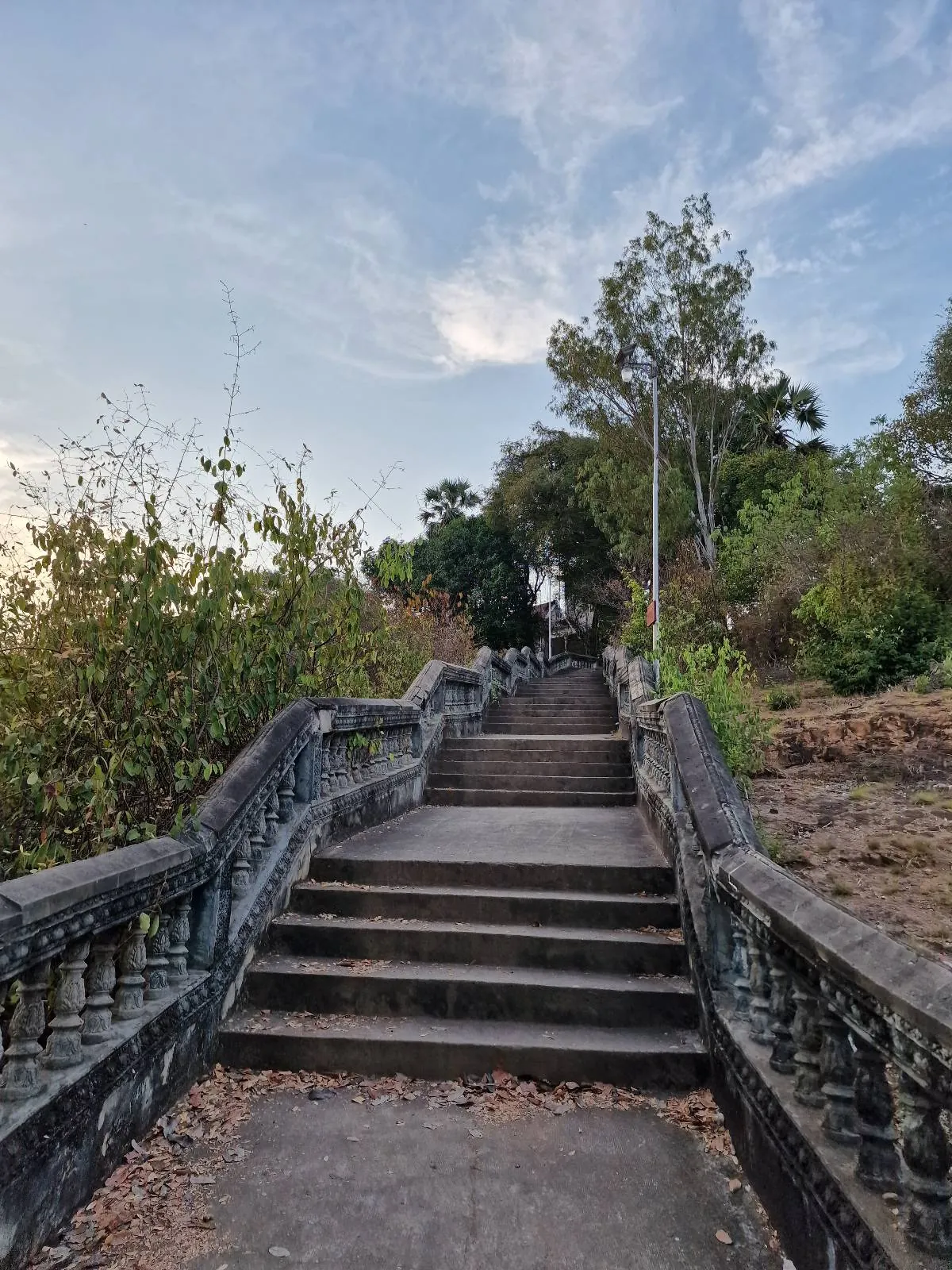 Stone steps with ornate railings lead upward through greenery and trees under a blue sky with scattered clouds. The pathway appears old and weathered, with leaves scattered on the steps.