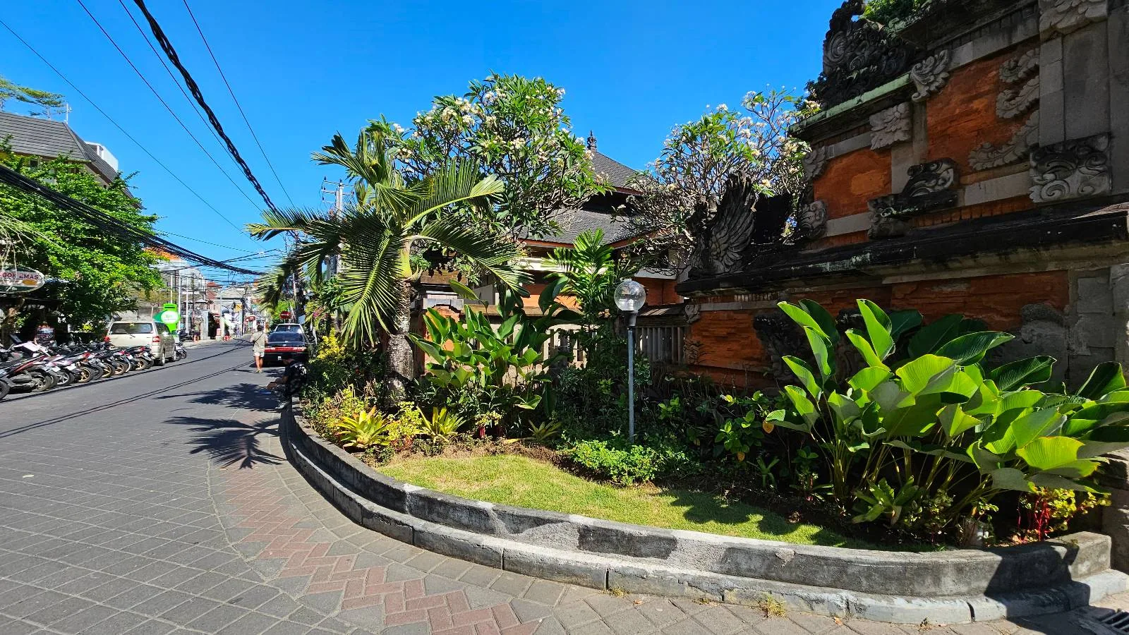 A narrow street curves to the left, bordered by vibrant green foliage and tropical plants. A traditional structure with ornate wooden details is partly visible. The sky is clear and blue, with motorcycles parked along the street.