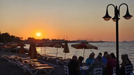 Sunset at Mavromatis Taverna in Greece, with beach umbrellas, sun loungers, and diners seated at tables beside the sea as the sky turns orange.