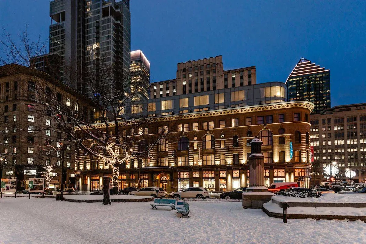 Snow-covered city square at dusk, with lit trees and historic building. Cars are parked, skyline towers above, creating a serene winter scene.