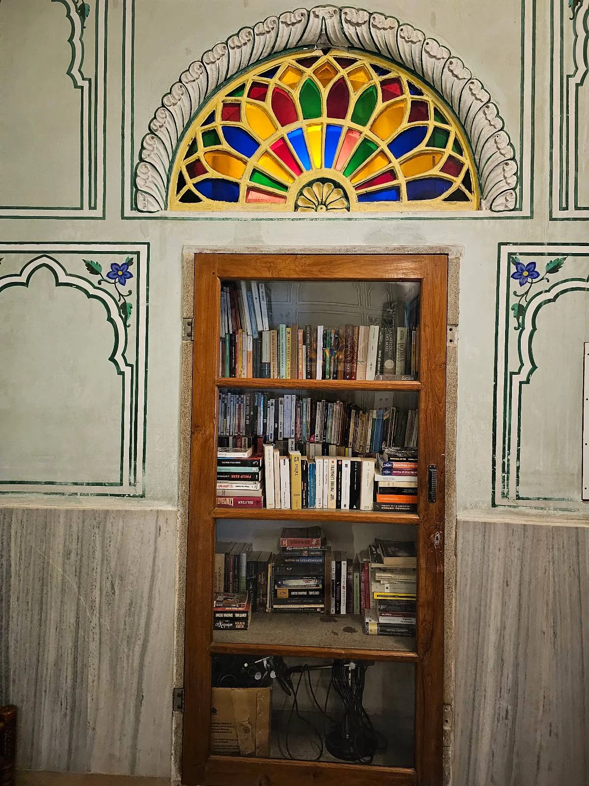 A wooden bookshelf filled with books stands against a decorated wall, topped by a colorful stained glass window shaped like a fan.