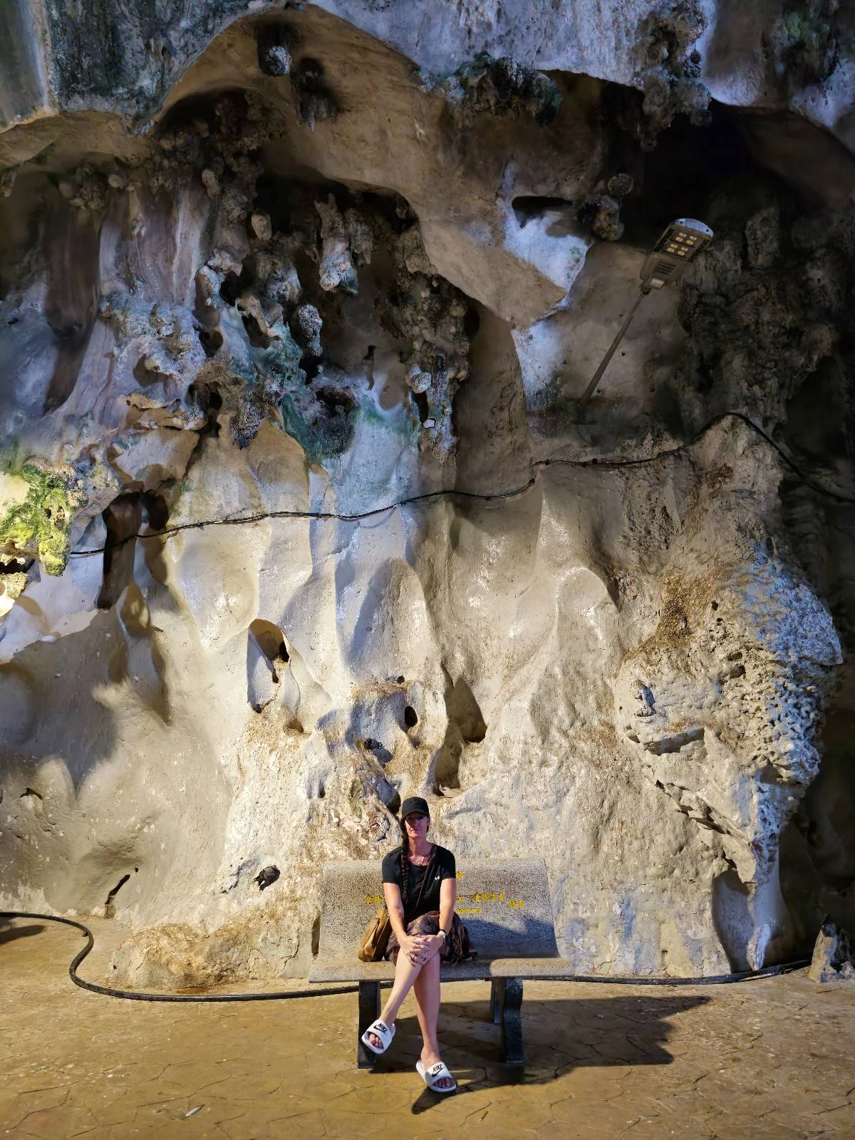 A person sits on a bench inside a cave with textured rock formations and stalactites overhead. The cave is dimly lit, highlighting the natural shapes and contours of the rocks. The person wears a dark shirt, shorts, and sneakers.