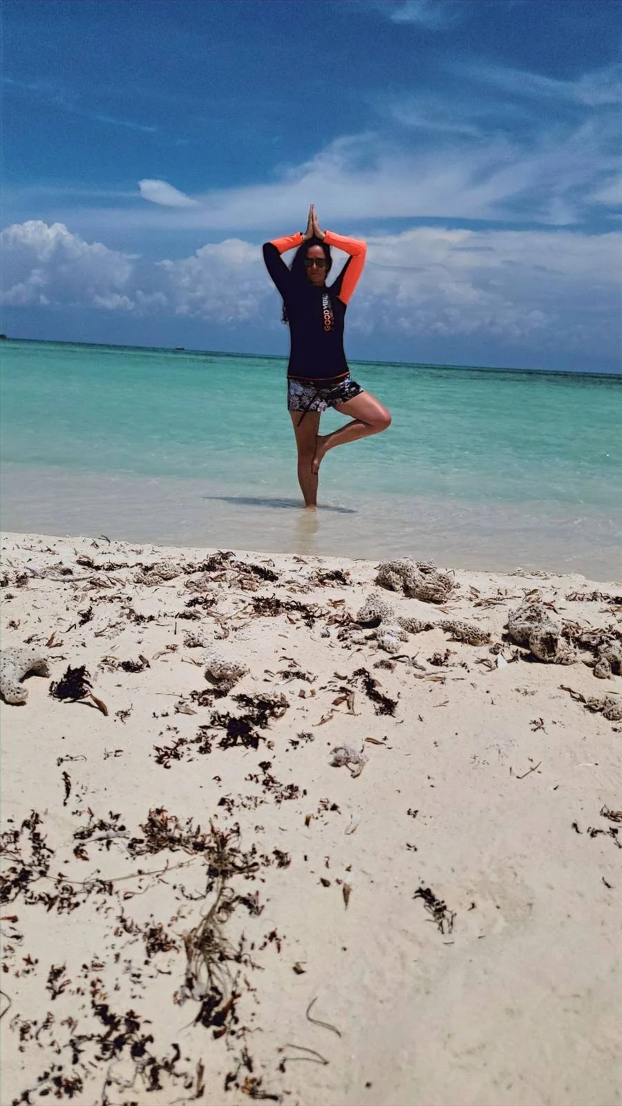 A person stands in a yoga pose on a sandy beach. The ocean is turquoise under a partly cloudy sky. Seaweed is scattered on the sand in the foreground.