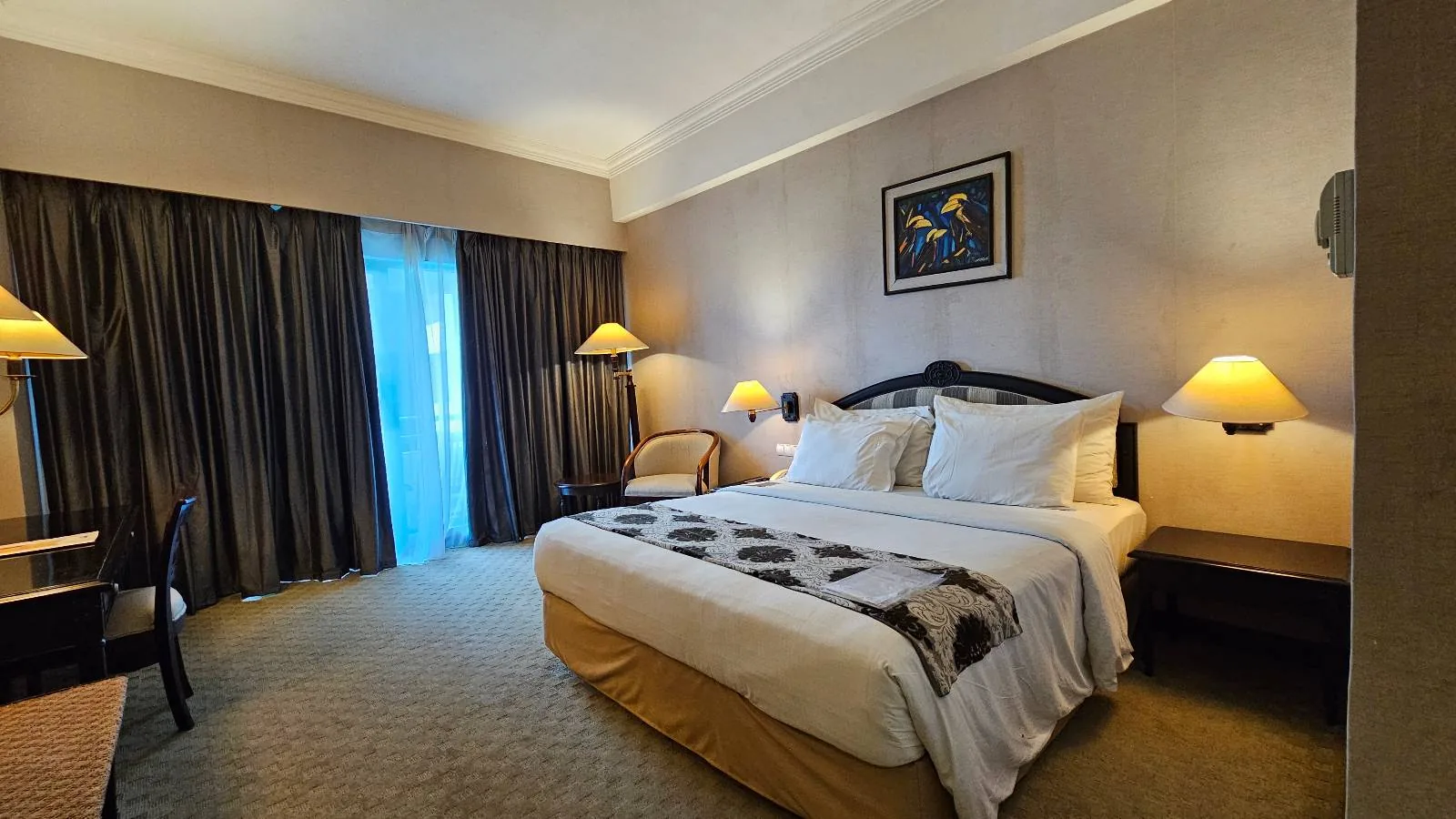 A well-lit hotel room with a large bed, neatly made and adorned with decorative pillows and a throw blanket. Two matching bedside tables with lamps flank the bed. A chair is in the corner, and a large window with curtains allows natural light in.