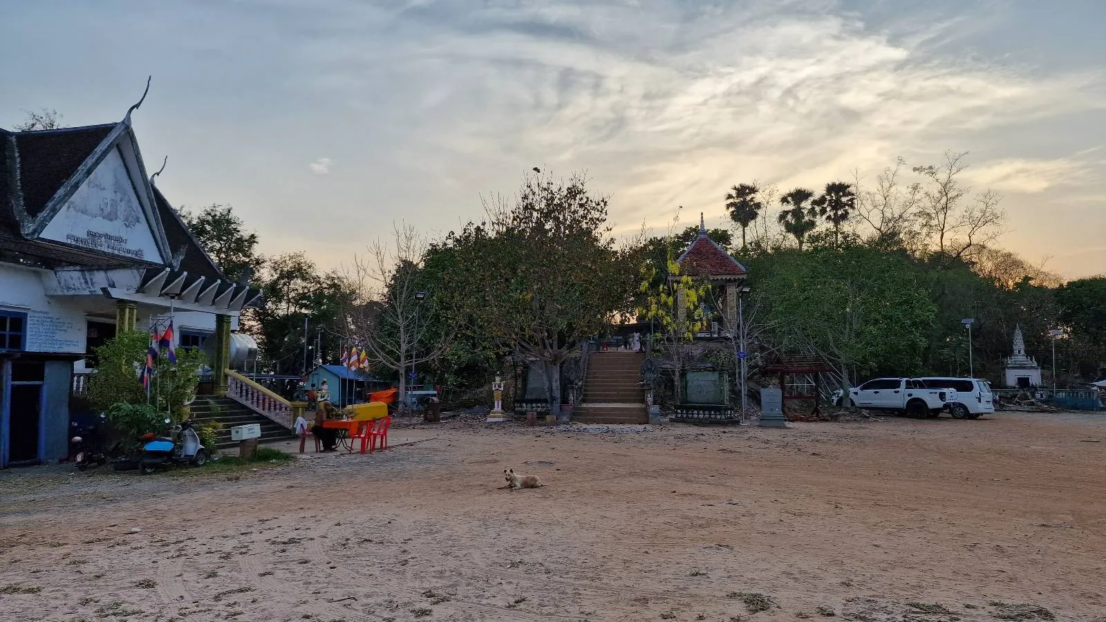 A traditional temple building with yellow and white walls, ornate roof decorations, and two staircases leading up to the entrance, set against a blue sky with trees in the background.