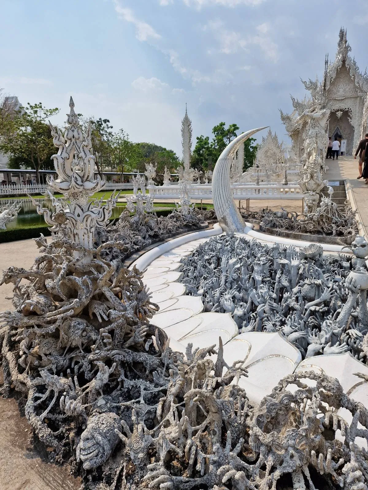 An ornate white sculpture garden with intricate, swirled designs and a crescent moon shape in front of a white temple under a partly cloudy sky.