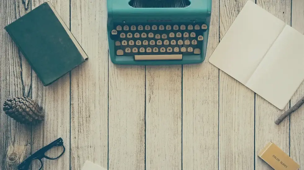 A vintage typewriter, book, open notebook, pinecone, and glasses arranged on a light wooden table with ample empty space in the center.