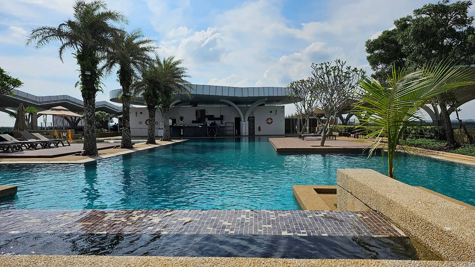 A serene swimming pool with clear blue water is surrounded by palm trees and lounge chairs under a partly cloudy sky. A modern building with a curved roof is visible in the background, adding a stylish touch to the tropical setting.