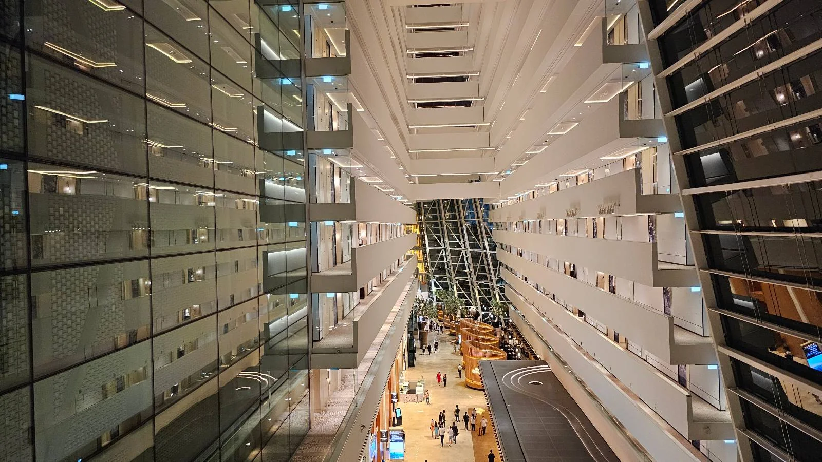 Looking down into a modern, spacious atrium with multiple floors. Glass walls and balconies line the sides. People and seating areas are visible on the ground floor. The architecture features symmetrical, geometric designs and a high ceiling.