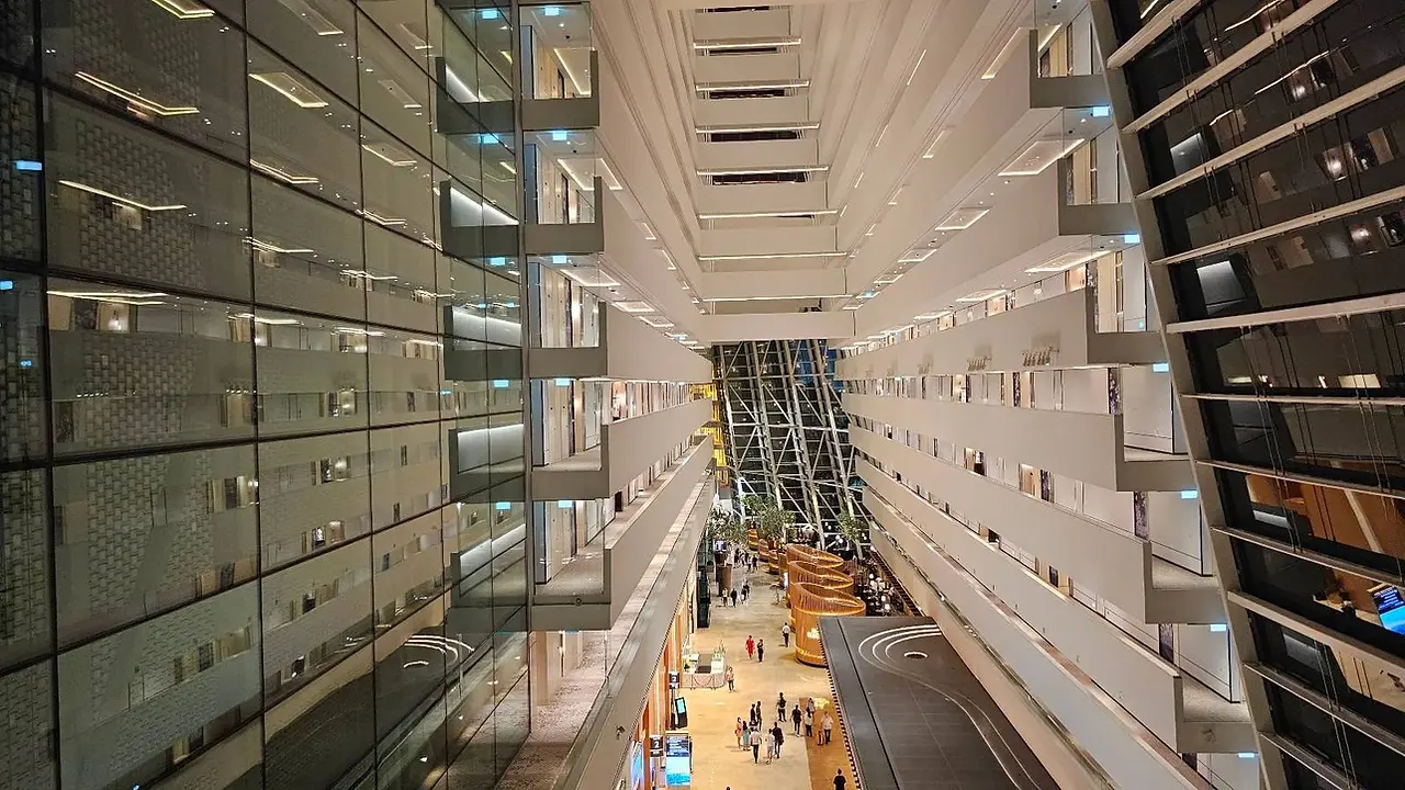 Looking down into a modern, spacious atrium with multiple floors. Glass walls and balconies line the sides. People and seating areas are visible on the ground floor. The architecture features symmetrical, geometric designs and a high ceiling.