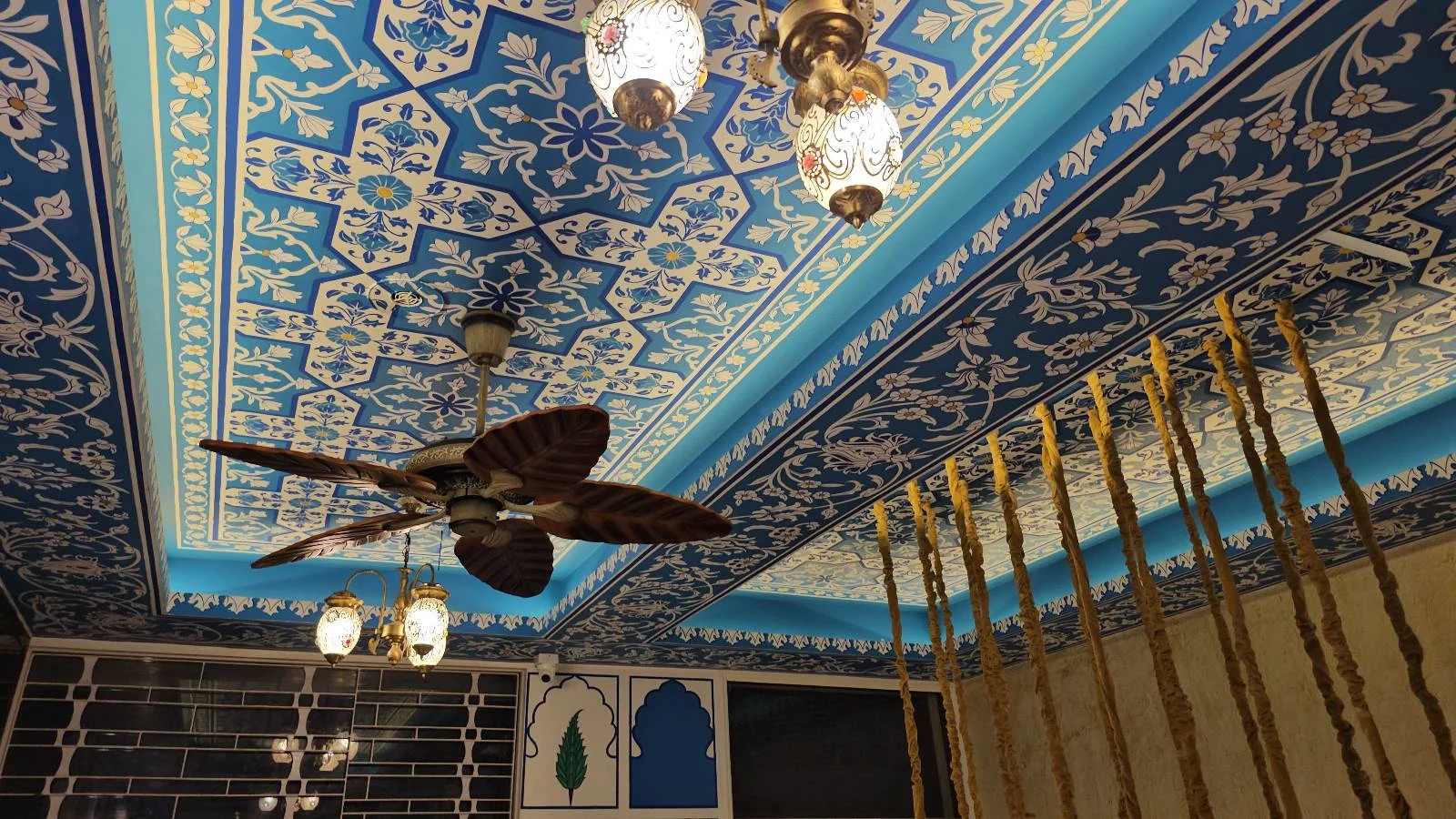 An ornate ceiling with intricate blue and white patterns, hanging lanterns, a decorative ceiling fan, and golden vertical rods. The design features traditional and elegant elements.