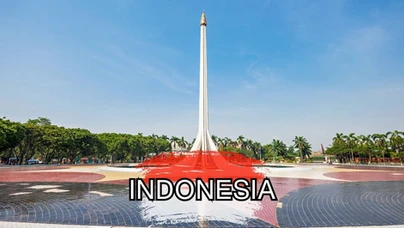 A tall white monument with a golden tip stands in a circular plaza surrounded by greenery under a clear blue sky. "INDONESIA" is written in bold letters over a red paint splash in the foreground.