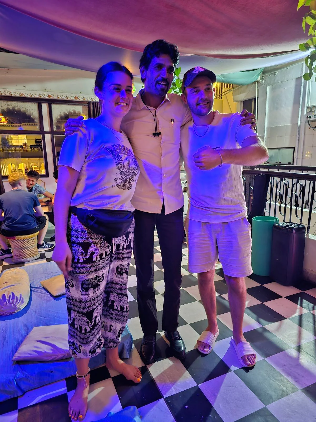 Three people pose together indoors on a black and white checkered floor. Two wear casual clothes, and one in the middle wears a white shirt and black pants. The scene is brightly lit with purple and blue lighting.