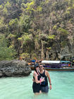 A couple stands in clear turquoise water near a rocky shore, embodying the spirit of slow travel. Smiling and embracing, with a wooden boat and lush greenery behind them, the woman dons sunglasses and a hat under the bright sun—a perfect digital nomad's retreat.
