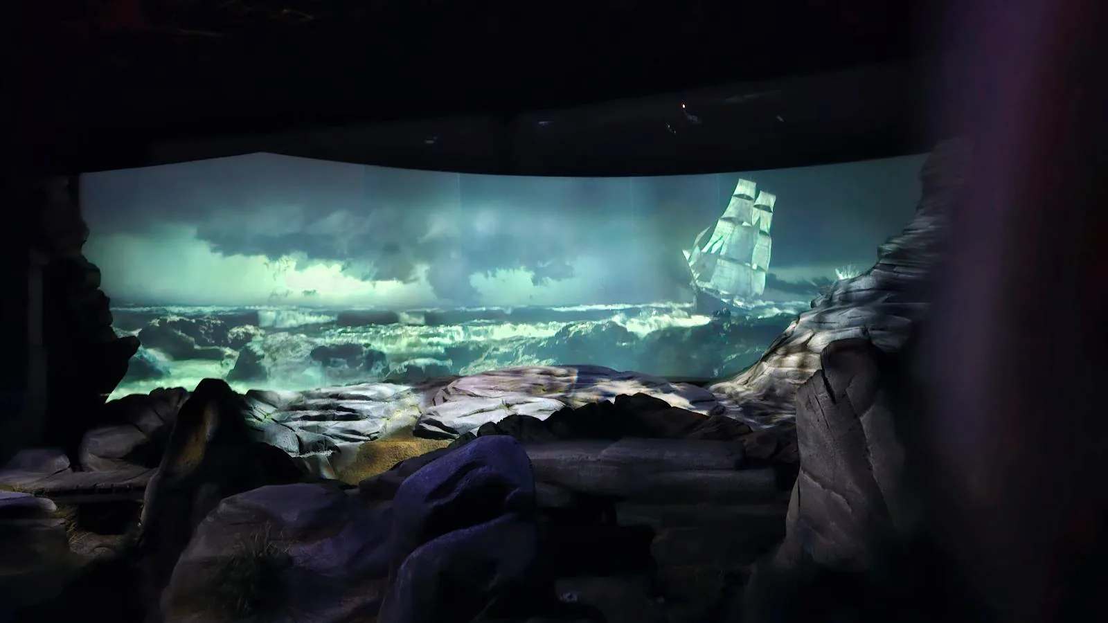 A dramatic scene of a tall ship sails through rough, stormy seas under a dark, cloudy sky, with rocky terrain and crashing waves in the foreground.
