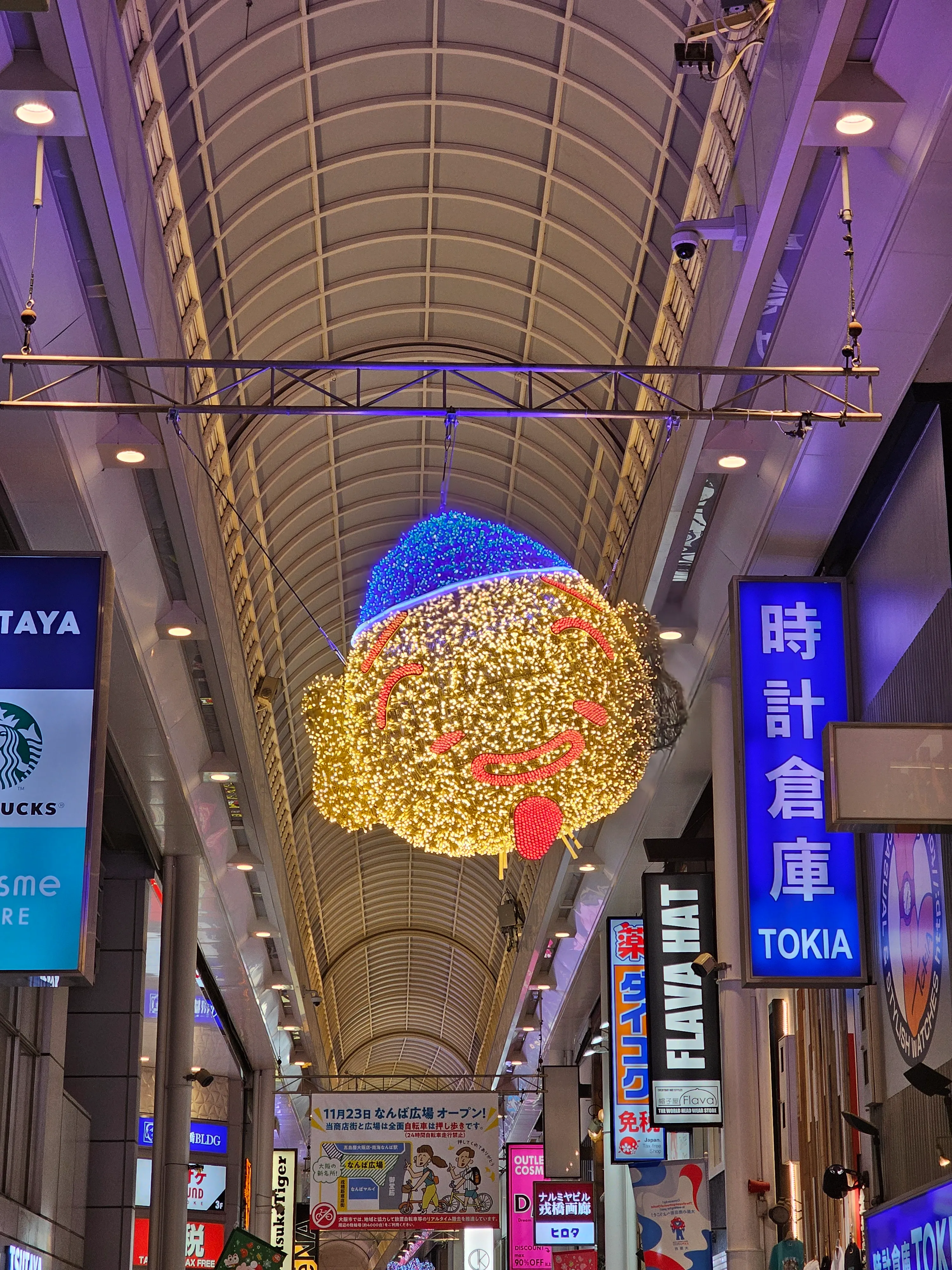A large, illuminated pufferfish decoration hangs from the ceiling of a brightly lit shopping arcade lined with colorful signs and storefronts.