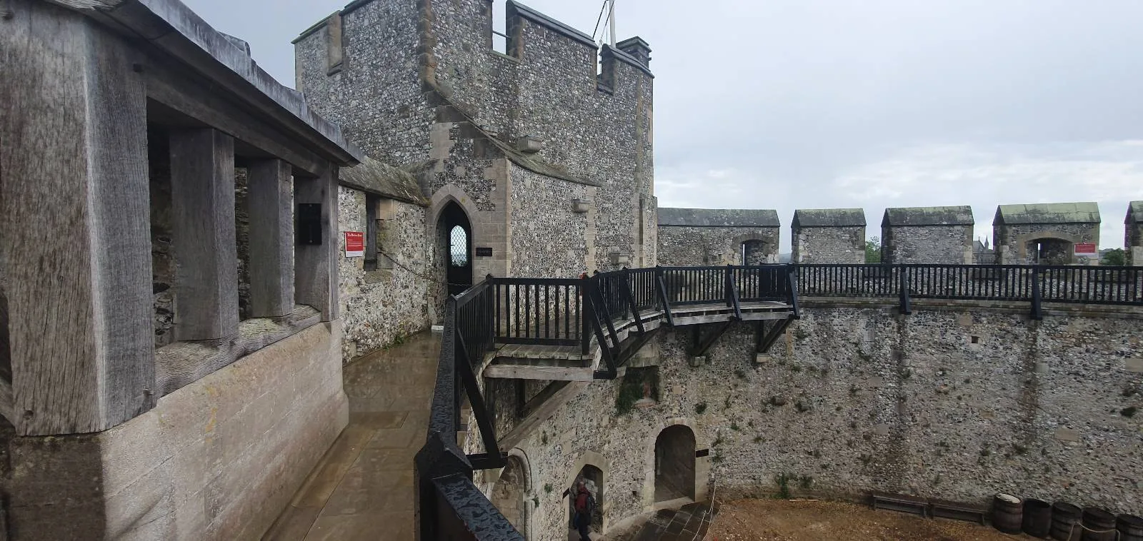 Stone interior of an ancient fortress with a round tower and battlements. A narrow wooden bridge and walkway span the inner circular courtyard. Overcast sky is visible in the background.