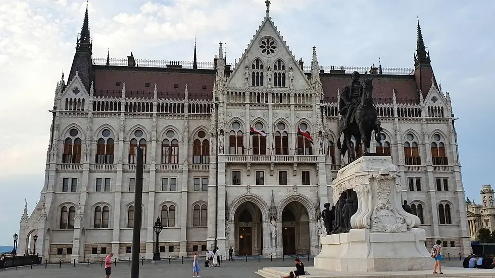 Historic building with arched windows and flags in the background. A statue of a rider on horseback in front. People walking nearby.