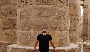 A man in a black t-shirt and light pants stands before a large ancient stone column with Egyptian hieroglyphs, looking up in awe. The background reveals more carved columns, capturing the wonder of Egypt travel.
