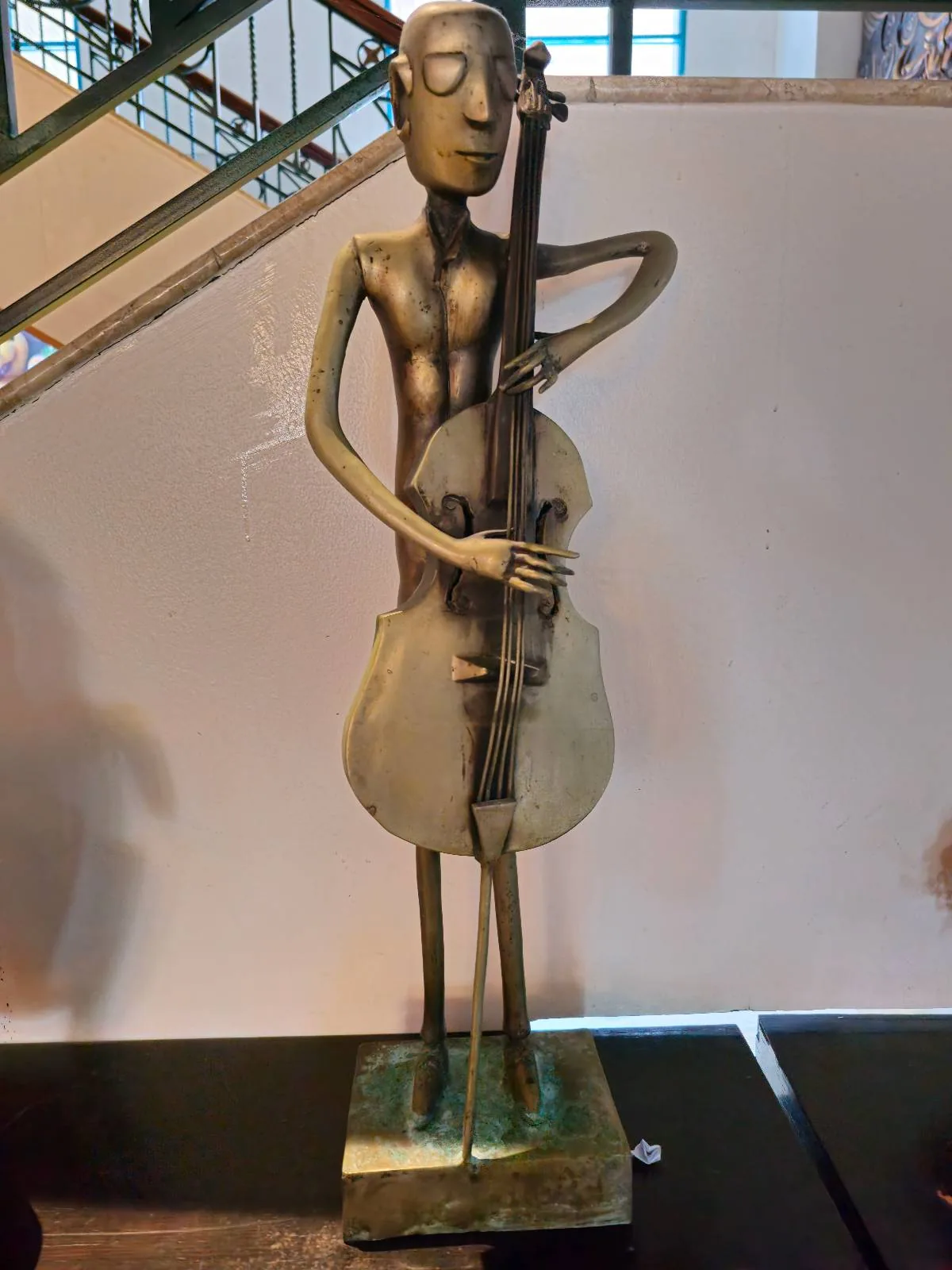 A tall, thin sculpture of a person playing a cello. The figure has elongated limbs and a minimalist design. The sculpture is placed against a light background with a staircase visible in the upper left corner.