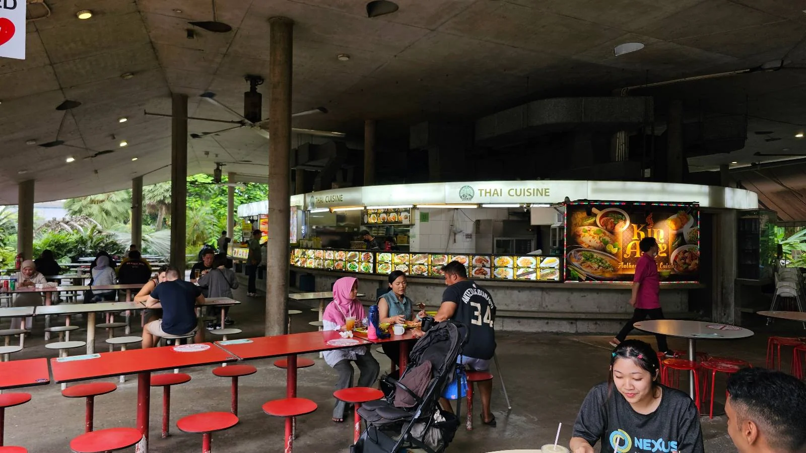 People are sitting and dining at a bustling food court with a round food stall in the background. The area is covered, with red benches and greenery visible outside.