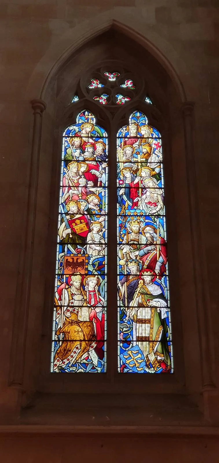 A tall, arched stained glass window in a church, featuring vibrant depictions of religious figures and scenes. The intricate designs in red, blue, yellow, and white are illuminated by natural light, casting colorful reflections on the interior's stone walls.