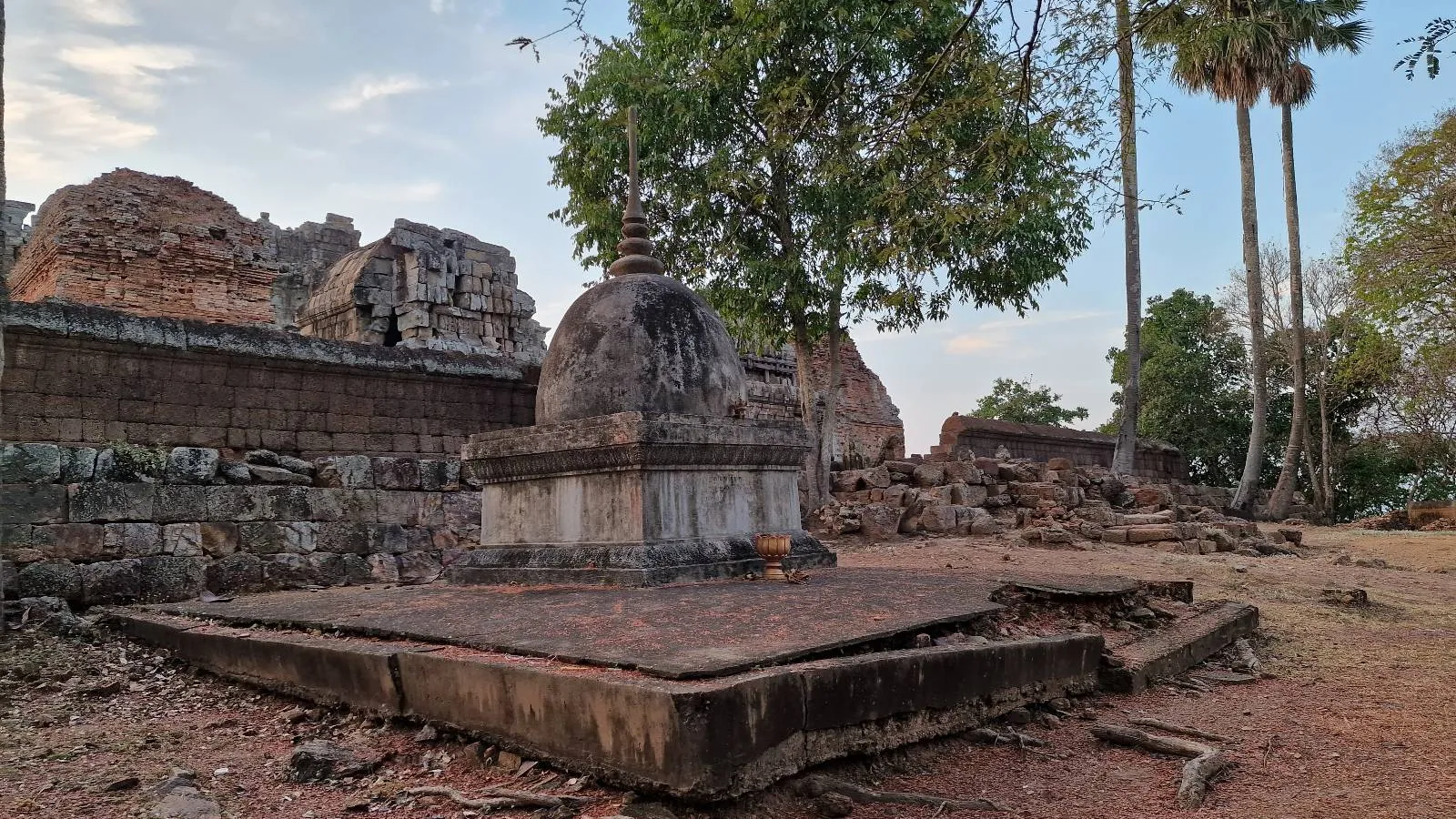 Ancient stone ruins with a weathered dome structure in the foreground, surrounded by scattered stones, trees, and remnants of old walls under a partly cloudy sky.