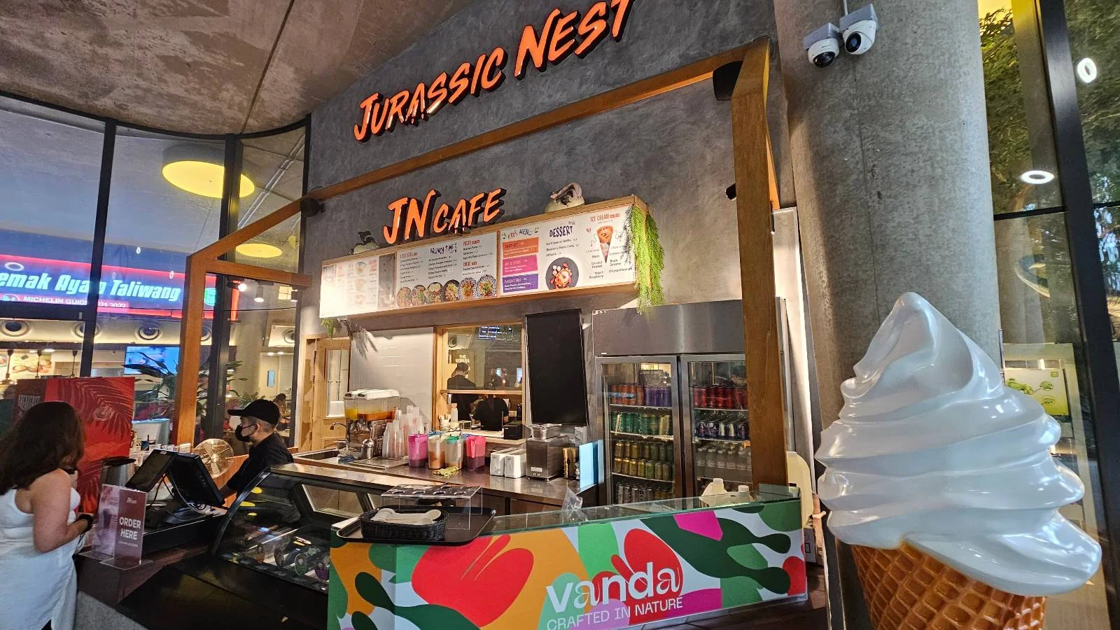 A cozy café called "Jurassic Nest JN Cafe" features a colorful counter decorated with vibrant designs. A large soft-serve ice cream cone display stands in the foreground, and a person is seen working behind the counter.