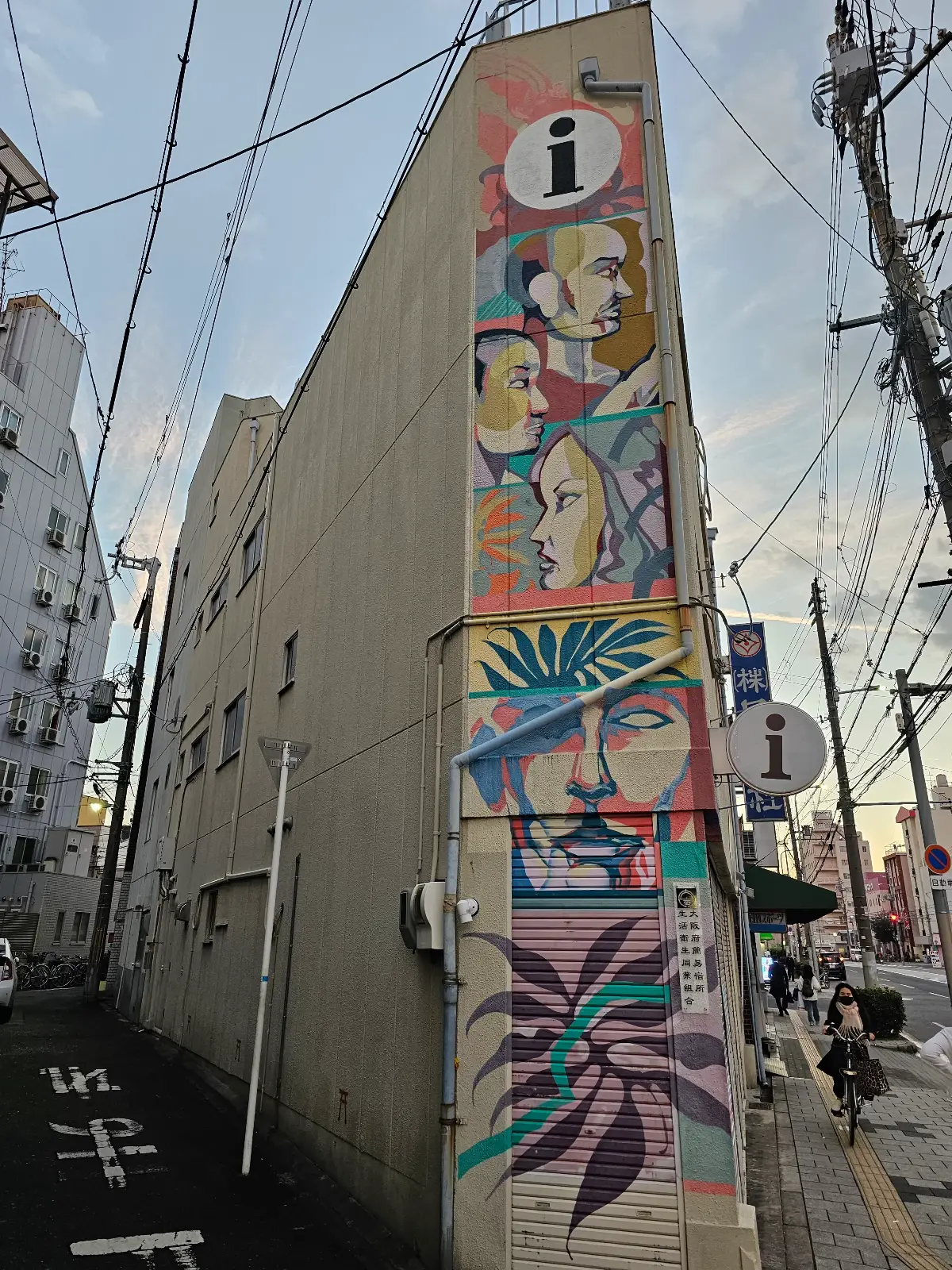 A colorful mural featuring abstract faces, geometric shapes, plants, and bold lines decorates the side of a narrow building on an urban street with power lines overhead.