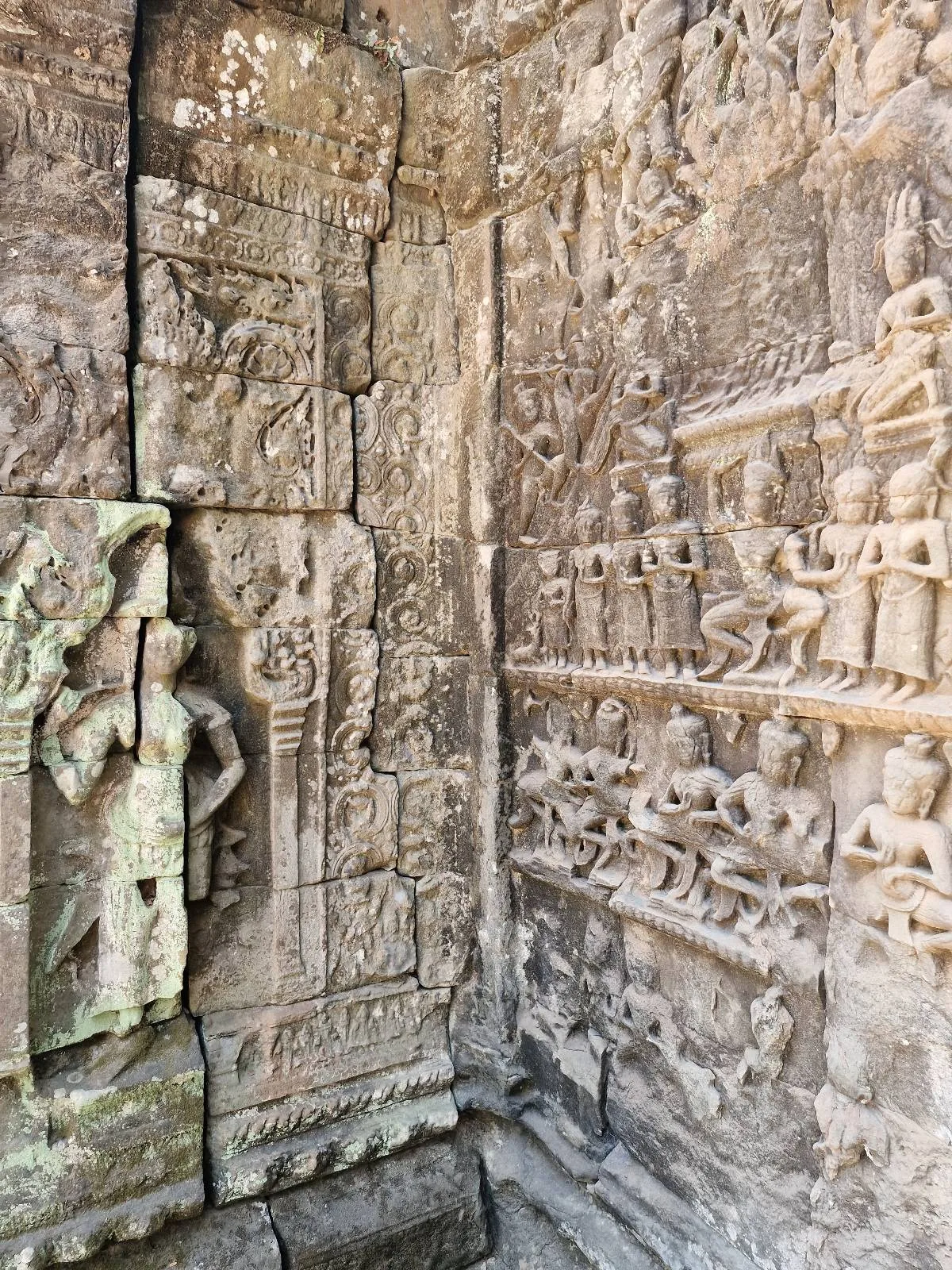 Ancient stone wall with intricate carvings and bas-reliefs, including a standing figure on the left and detailed patterns and figures covering the surrounding surface.