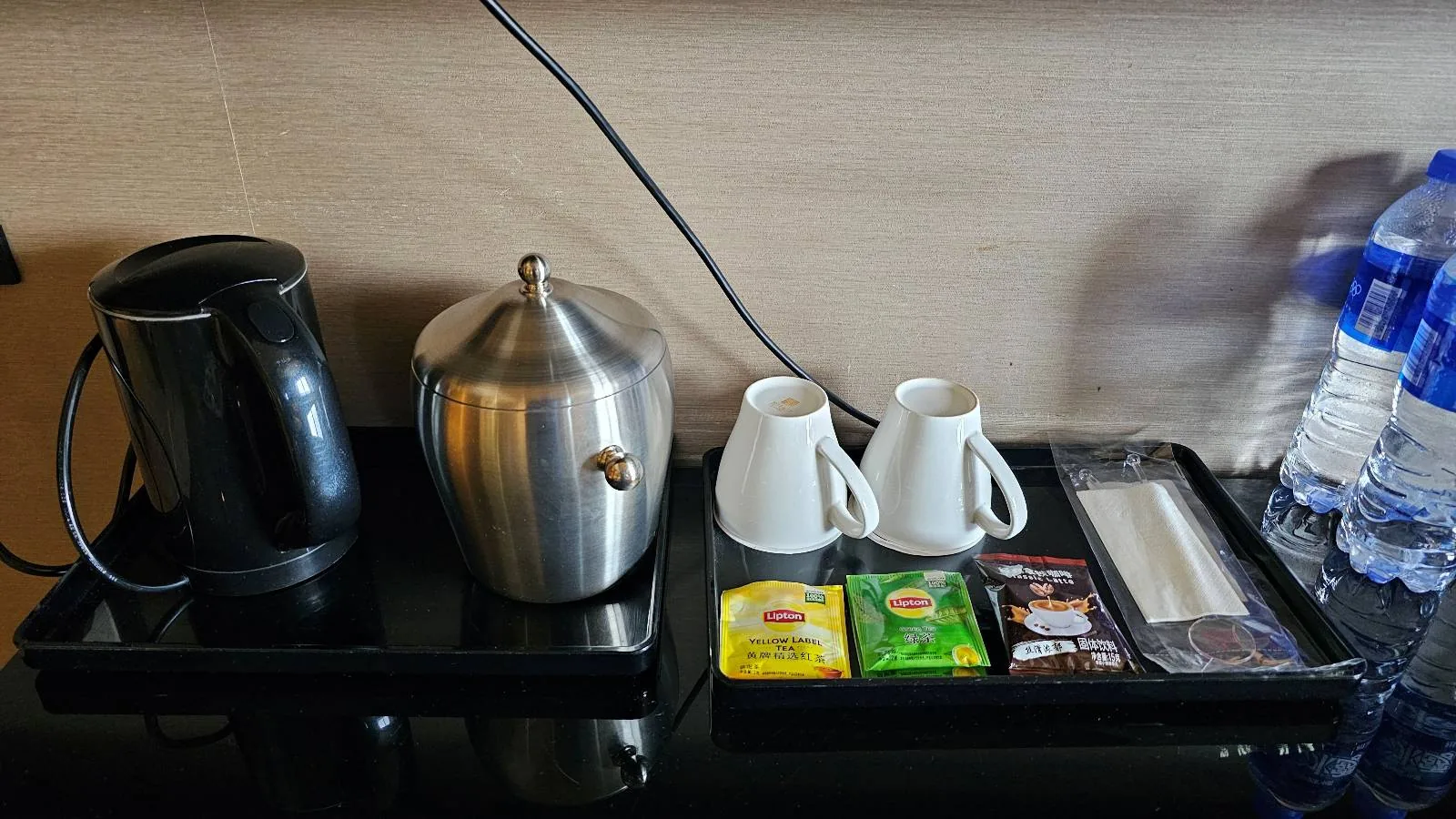 A hotel room beverage station with an electric kettle, an ice bucket, two white cups, tea and coffee sachets, stirrers, and two bottles of water arranged neatly on a black tray.