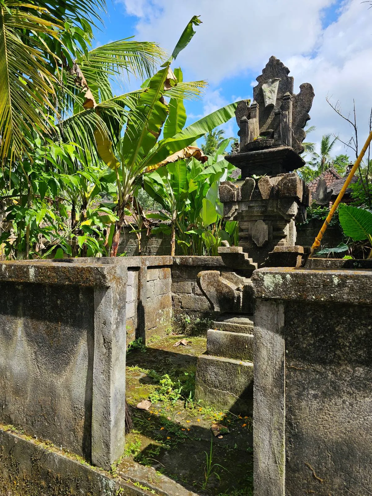 A stone shrine with intricate carvings is surrounded by a moss-covered stone wall. Lush green banana plants and palm trees are in the background under a partly cloudy sky.