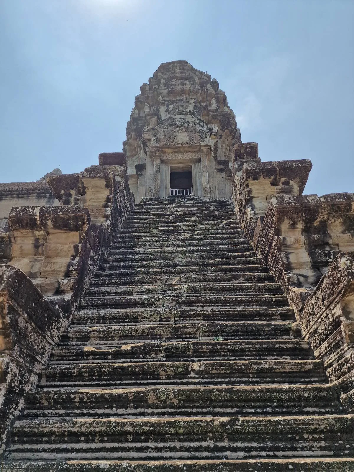Steep stone staircase leads up to an ancient temple structure with weathered carvings, set against a bright, partly cloudy sky.