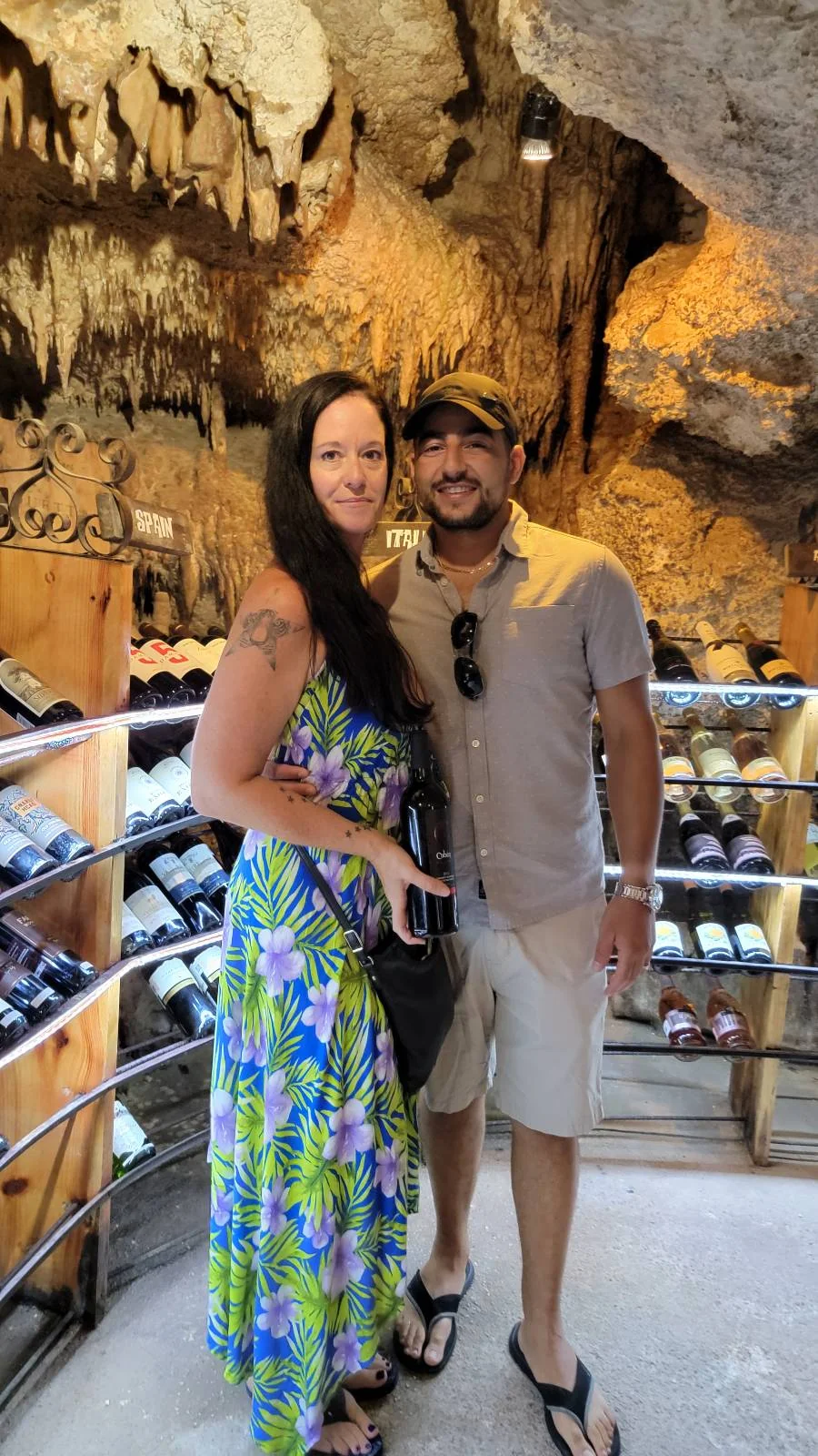 A couple stands in a wine cellar with rocky walls and a collection of wine bottles. The woman is wearing a floral dress and the man is in a light shirt and shorts. They are both smiling and standing close together.