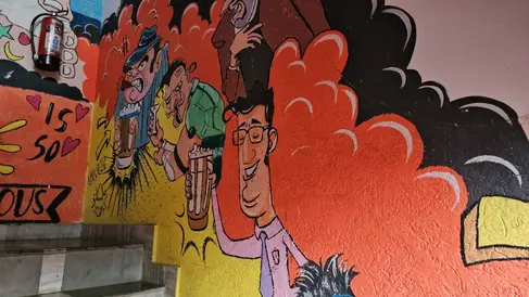A vibrant mural on a wall depicts cartoonish characters with exaggerated expressions against a colorful background of red, orange, and black swirls. Text and hearts are visible on the left side. A staircase is partially visible in the foreground.