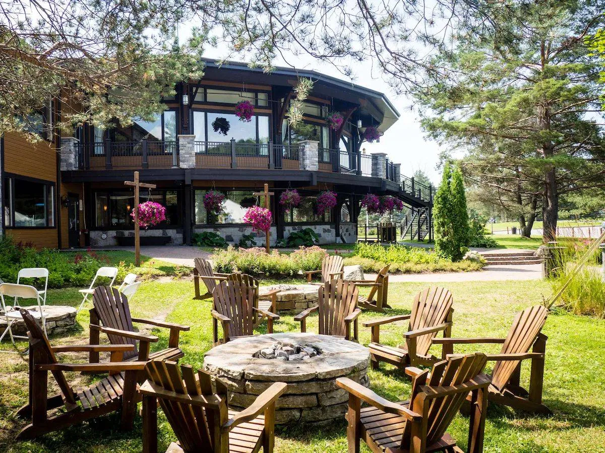 Wooden chairs encircle a stone fire pit in a garden. A large building with hanging baskets is in the background, surrounded by trees. Peaceful setting.