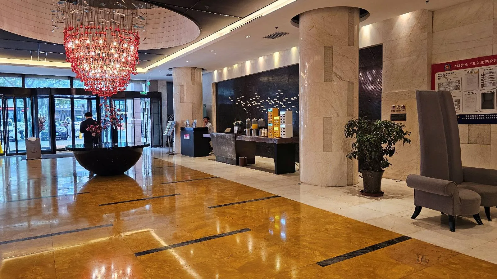 Modern hotel lobby with shiny marble floors, a large red chandelier, tall pillars, potted plants, and elegant seating. A reception desk is visible in the background near the glass entrance doors.