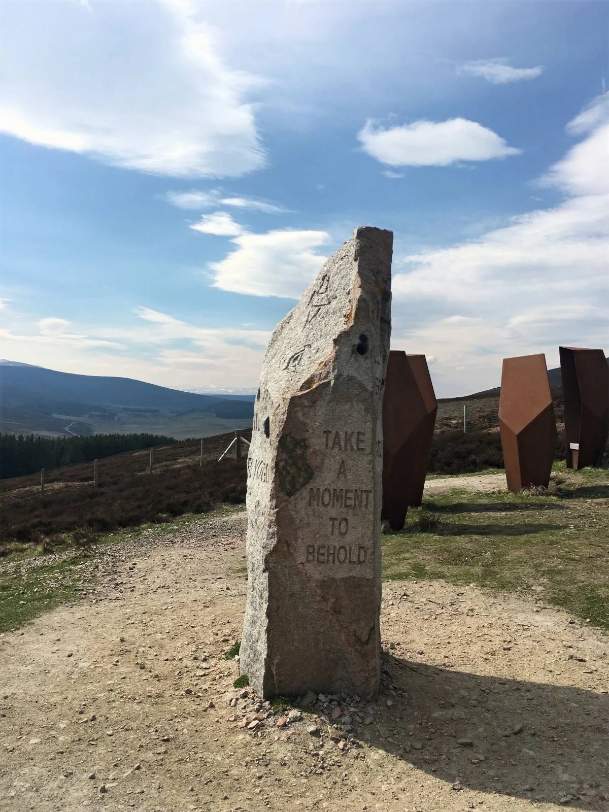 A large stone stands in a hilly landscape with a distant row of trees. The stone bears the inscription, "Take a moment to behold. As still skies or storms unfold." The sky is mostly clear with a few clouds.