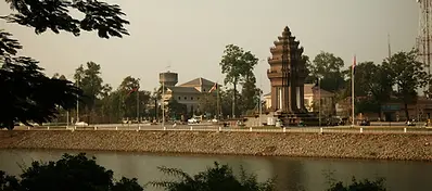 A view across a river in Battambang showing a brown stone monument, Cambodian flags, and city buildings under a hazy sky.