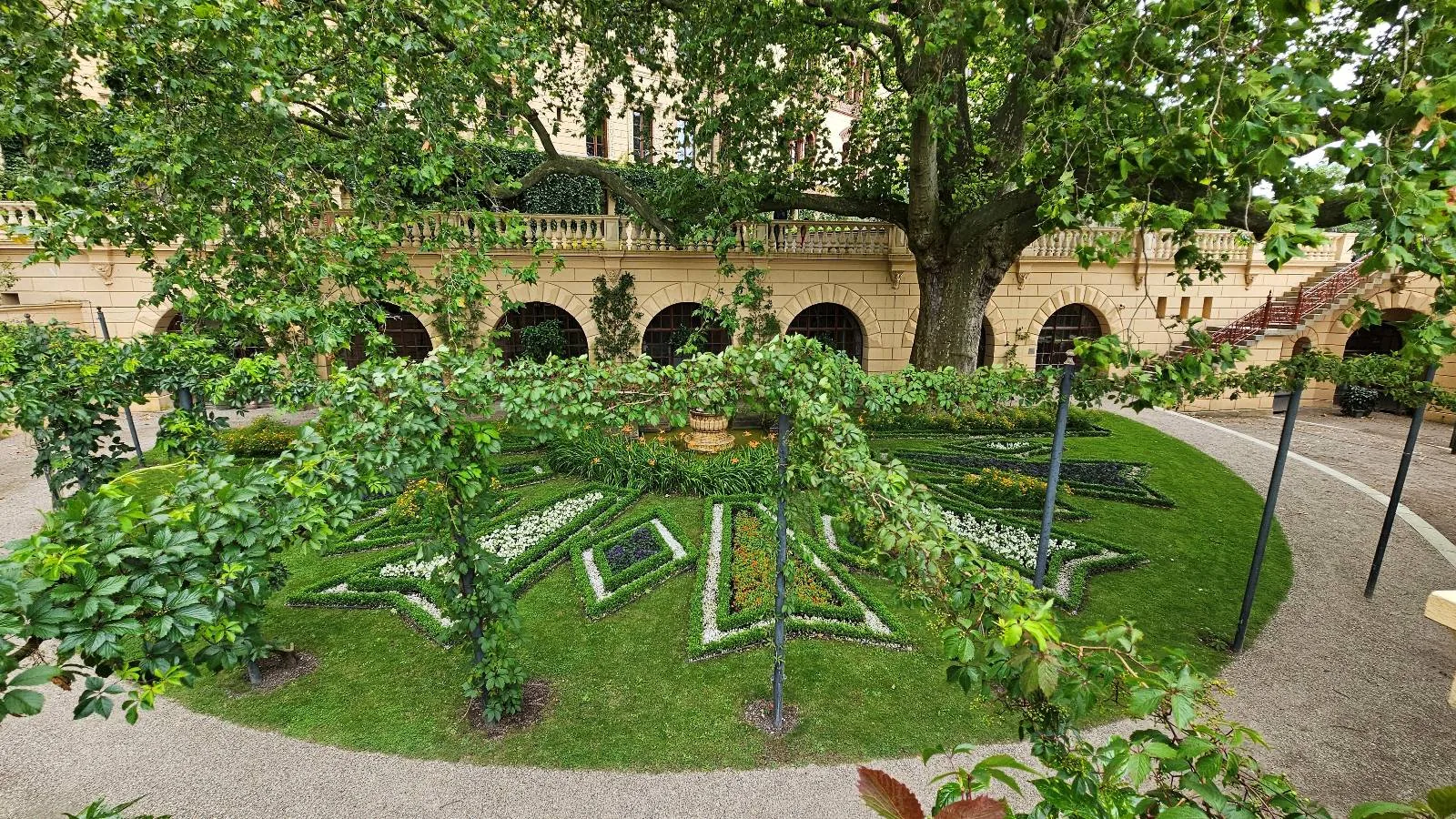 A lush garden with manicured lawns, symmetrical flower beds, and green archways, surrounded by a stone building with arches and shaded by large trees.