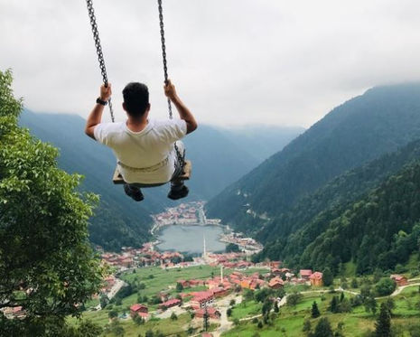 High above a lush green valley, with a breathtaking view of the village and mountain lake of Uzungöl in Trabzon Province, a person swings amidst the grandeur of forested mountains under a cloudy sky.