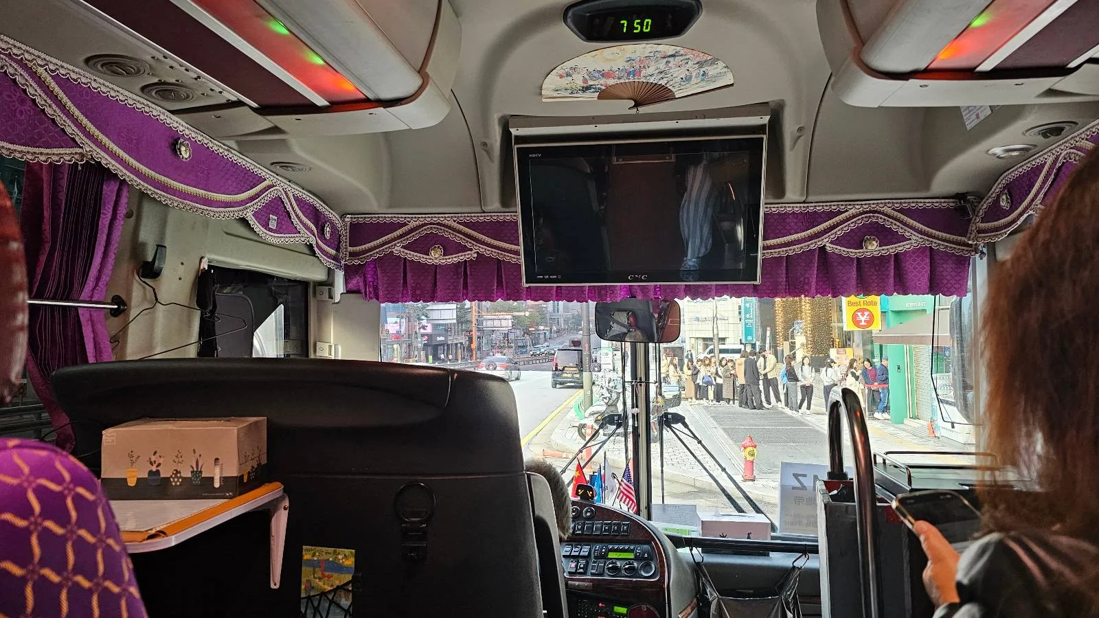 Interior view of a bus with a purple curtain, featuring a screen displaying a video. The driver is visible, and passengers can be seen through the front window. People are walking outside on a street.