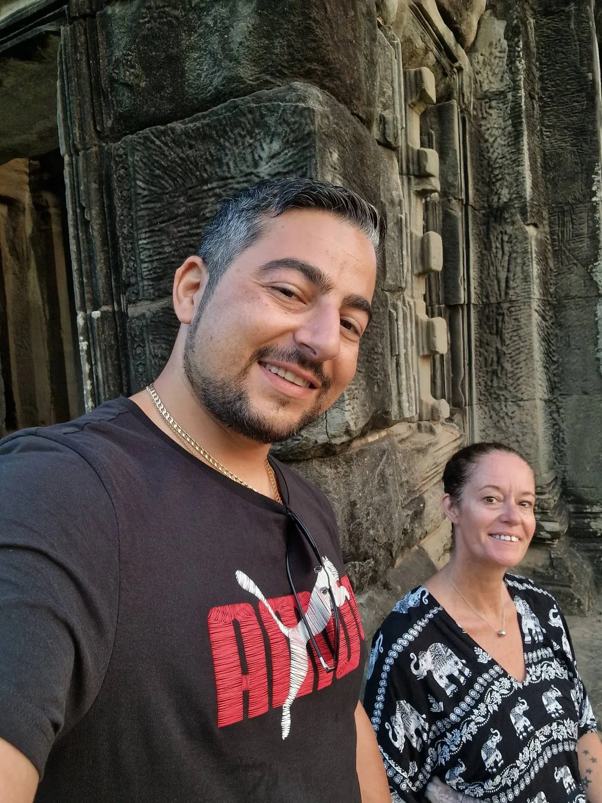 A man and a woman smile while taking a selfie in front of ancient stone ruins. The man wears a black Adidas shirt, and the woman wears a patterned black-and-white top. The background shows weathered stone walls.