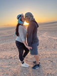 As the sun sets on the desert horizon, a couple, embodying the spirit of slow travel, stands hand in hand. They lean in for a kiss, headscarves fluttering gently. The warm glow envelops them, capturing their closer lives in this serene and timeless moment.
