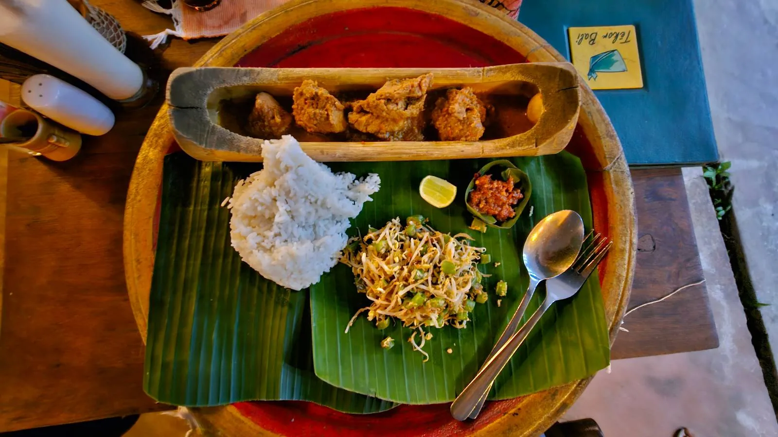 A plate of rice with shredded vegetables wrapped in banana leaves sits beside a small bowl of sauce and lime wedge. Fried dumplings are arranged in a row on a long dish. Cutlery is placed on the side. The table is set for a meal.